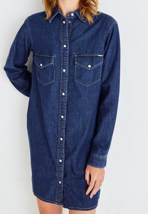 Femme portant une robe chemise en denim à manches longues avec des boutons pression sur le devant et deux poches poitrine, debout devant un fond blanc uni.