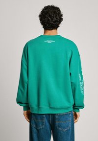 Teal sweatshirt van katoen, met een relaxed fit en geribbelde manchetten. Heeft logo-tekst op de achterkant en op de mouw. Gecombineerd met spijkerbroeken.
