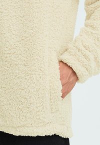 Cremefarbener Fleece-Pullover mit einer weichen, strukturierten Oberfläche. Verfügt über eine Tasche auf der rechten Seite und elastische Bündchen für eine enge Passform.