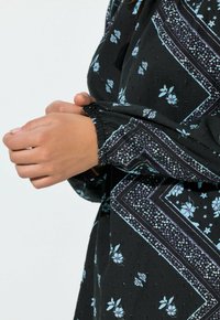 Blusa negra con estampado de flores en azul claro y púrpura, puños fruncidos y tejido texturizado con pequeños puntos.