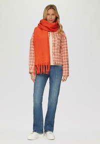 Écharpe orange avec des bords à franges, associée à une veste pied-de-poule, un pull léger et un jean bleu. Les chaussures comprennent des baskets blanches à plateforme.