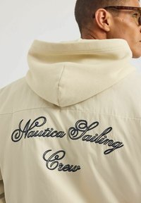 Beżowa kurtka z miękkim, kapturowym kołnierzem. Zawiera haftowany napis "Nautica Sailing Crew" w ciemnoniebieskim kolorze z tyłu. Gładka tekstura.
