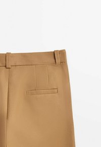 Ljusbruna byxor visas, med midjeband, bältesöglor och en bakficka, mot en slät vit bakgrund.