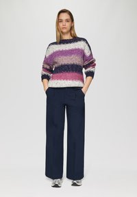 Gehaakte trui met een meerkleurig patroon in paars, wit en marineblauw; gecombineerd met marineblauwe wijde broek; model heeft haar handen in de zakken.