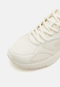 Hvit atletisk sneakers som har en mesh-overdel, polstret såle og kontrasterende teksturerte detaljer. Snøringen er lys og flat.