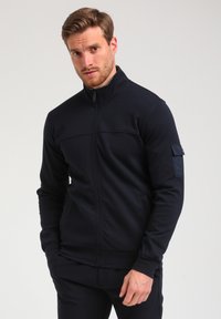 Veste zippée bleue marine en tissu doux et extensible. Présente un col haut, des détails de couture horizontale et une petite poche à badge sur la manche.
