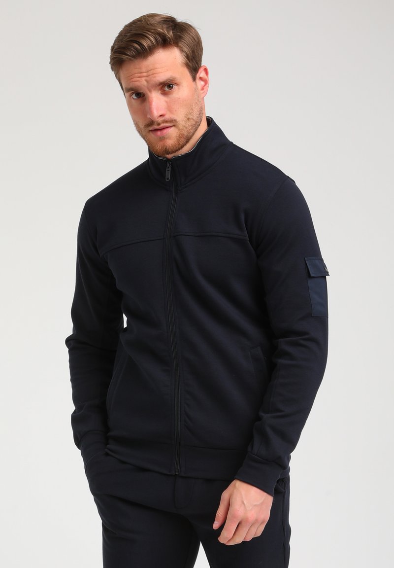 Veste zippée bleue marine en tissu doux et extensible. Présente un col haut, des détails de couture horizontale et une petite poche à badge sur la manche.