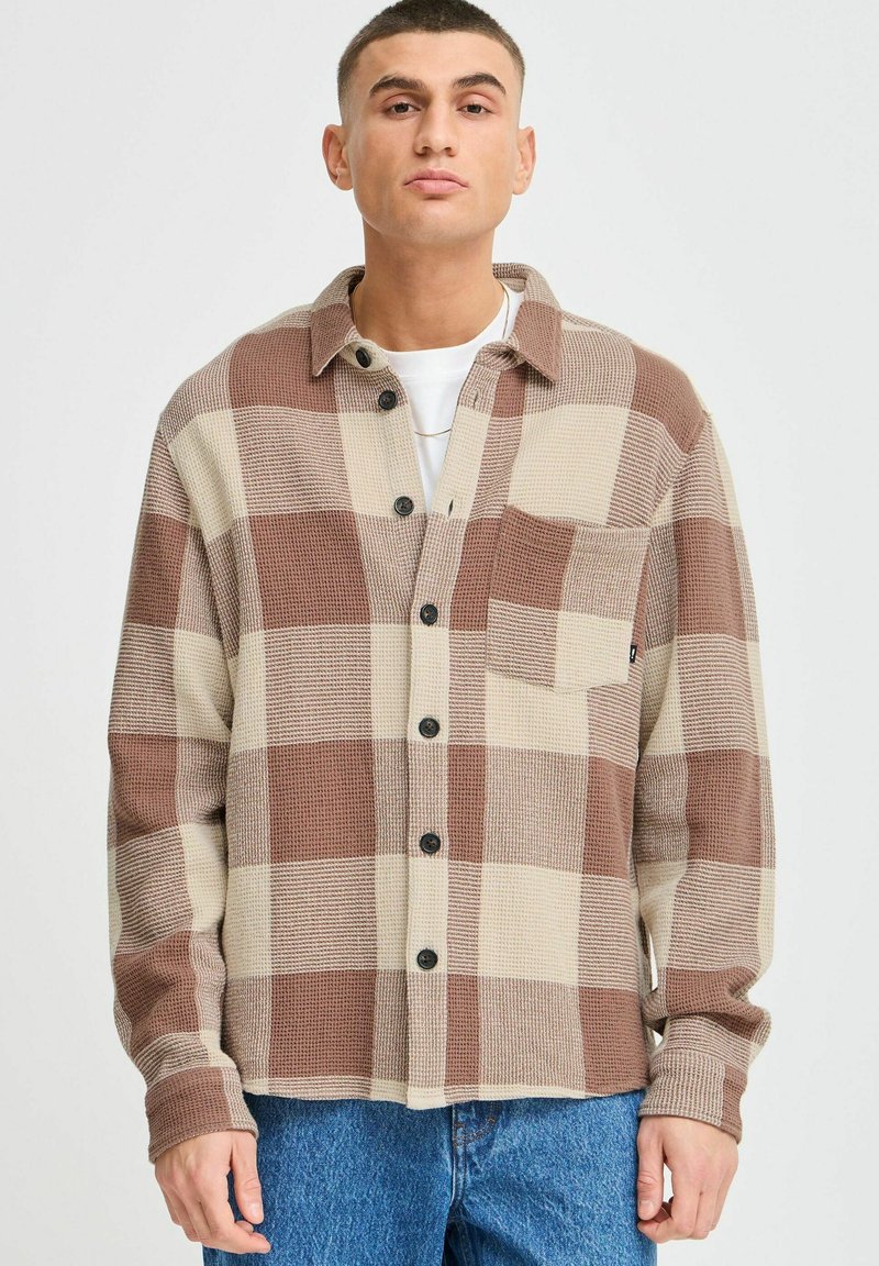 Brown and beige plaid button-up shirt with a chest pocket, made of textured fabric. Features black buttons and a classic collar.