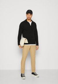 Black knitted pullover with a collar, beige trousers, and black high-top sneakers. Holding a light-colored jacket. White background.