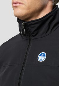 Schwarze Jacke mit hohem Kragen, ausgestattet mit einem Frontreißverschluss und einem Metallring zum Ziehen. Blau-weißes "North Sails"-Logo im Brustbereich.