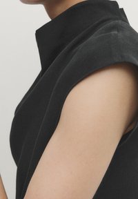 Black sleeveless garment with a high collar, featuring a structured design and a smooth texture, highlighting the shoulder area and neckline.