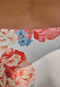 Látka s kvetinovým vzorom, na ktorej sú veľké červené a modré kvety na bielom pozadí; na vrchu sa nachádza malý zlatý kovový detail.