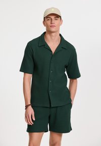 Cămașă verde texturată cu mânecă scurtă și închidere cu buton, având guler și pantaloni scurți asortați. Băciță bej cu un accent mic al logo-ului.
