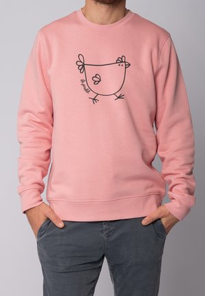 Mann trägt ein rosa Sweatshirt mit einer einfachen schwarzen Linienzeichnung eines Huhns und dem Text "Le poulet" auf der Vorderseite, die Hände in den Taschen einer grauen Hose.