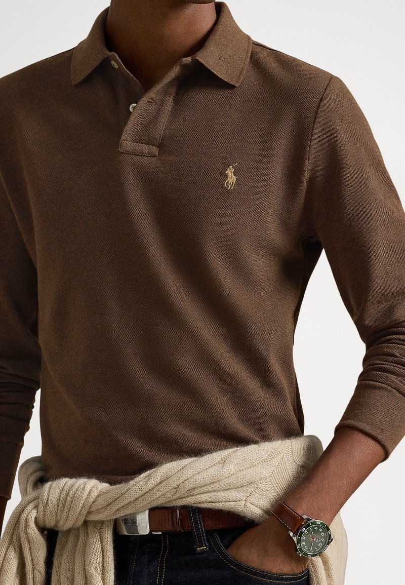 Braunes Langarm-Poloshirt mit strukturiertem Stoff und einem goldenen Logo auf der Brust. Kombiniert mit dunklen Jeans und einem cremefarbenen Strickpullover, der in der Taille gebunden ist.