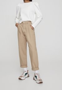 Pantalones beige de tiro alto con corte relajado y puños enrollados, combinados con una blusa blanca de manga larga con hombros abullonados y zapatillas.