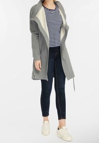 Grau langer Cardigan mit cremefarbener Fütterung, kombiniert mit einem navy- und weiß gestreiften Hemd und dunklen Jeans. Verfügt über eine lockere Passform und Reißverschlüsse.