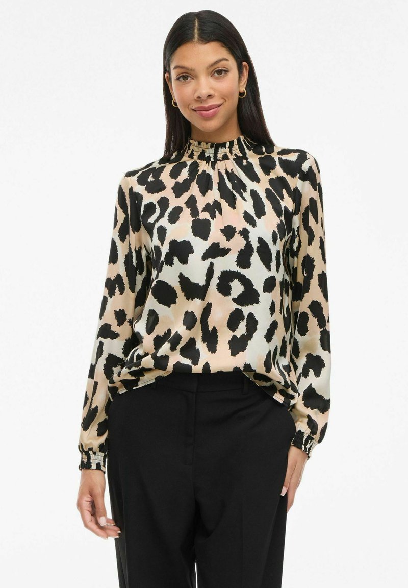 Woman wearing a beige and black leopard print long-sleeve blouse with ruffled collar and black high-waisted pants, smiling and standing.