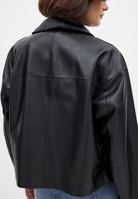 Juodas odinis švarkas su apykakle, šiek tiek sutrumpinta forma, lygia tekstūra ir matomais siūlių detalėmis nugaroje.