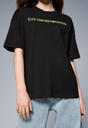Personne portant un t-shirt noir EA7 Emporio Armani avec un logo vert et un jean bleu clair sur un fond gris.