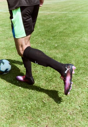 Des chaussures de football noires et roses avec un motif dégradé, portées avec des chaussettes montantes noires et un short noir avec des accents verts et bleus, sur de l'herbe.