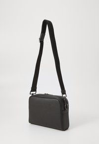 Sac bandoulière noir en matériau texturé, de forme rectangulaire, avec fermeture éclair et bandoulière ajustable ornée de logos.