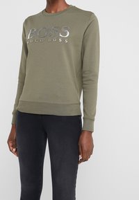 Persona che indossa una felpa color verde oliva di Hugo Boss con logo argentato e pantaloni neri attillati, su uno sfondo bianco semplice.