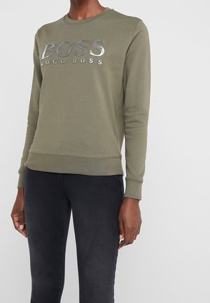 Persona che indossa una felpa color verde oliva di Hugo Boss con logo argentato e pantaloni neri attillati, su uno sfondo bianco semplice.