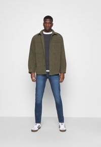 Olive green textured jacket over a gray sweater, paired with blue jeans and white sneakers with black stripes. Two breast pockets on the jacket.