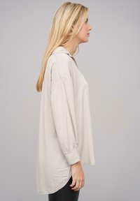 Camicia a maniche lunghe beige chiaro con un fit rilassato, scollatura rotonda e leggeri arricciamenti ai polsini. Texture morbida con un orlo curvo.