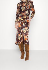 Robe à imprimé floral sur fond marron, corsage froncé, encolure à volants, bottes en suède marron montant jusqu'aux genoux, et une fente latérale dans la jupe.