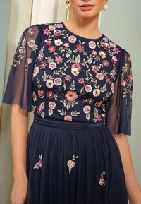 Dunkelblaues Kleid mit kurzen, transparenten Ärmeln, das mit floralen Stickereien in Rosa und Creme verziert ist. Fließender Rock mit zusätzlichen floralen Details.