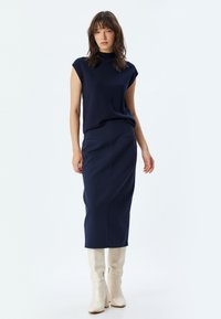 Marineblaue ärmellose Bluse kombiniert mit einem figurbetonten marineblauen Midirock. Das Modell trägt cremefarbene kniehohe Stiefel. Glatter Stoff mit einfachem Design, keine sichtbaren Muster.