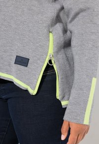 Sweatshirt gris avec une fermeture éclair jaune et des accents. Il présente une glissière sur le côté, des poignets côtelés et une étiquette de marque rectangulaire.