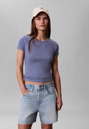 Kobieta ubrana w dopasowaną lawendową bluzkę z krótkim rękawem, jasne szorty dżinsowe i beżową czapkę z daszkiem, stojąca na jednolitym szarym tle.