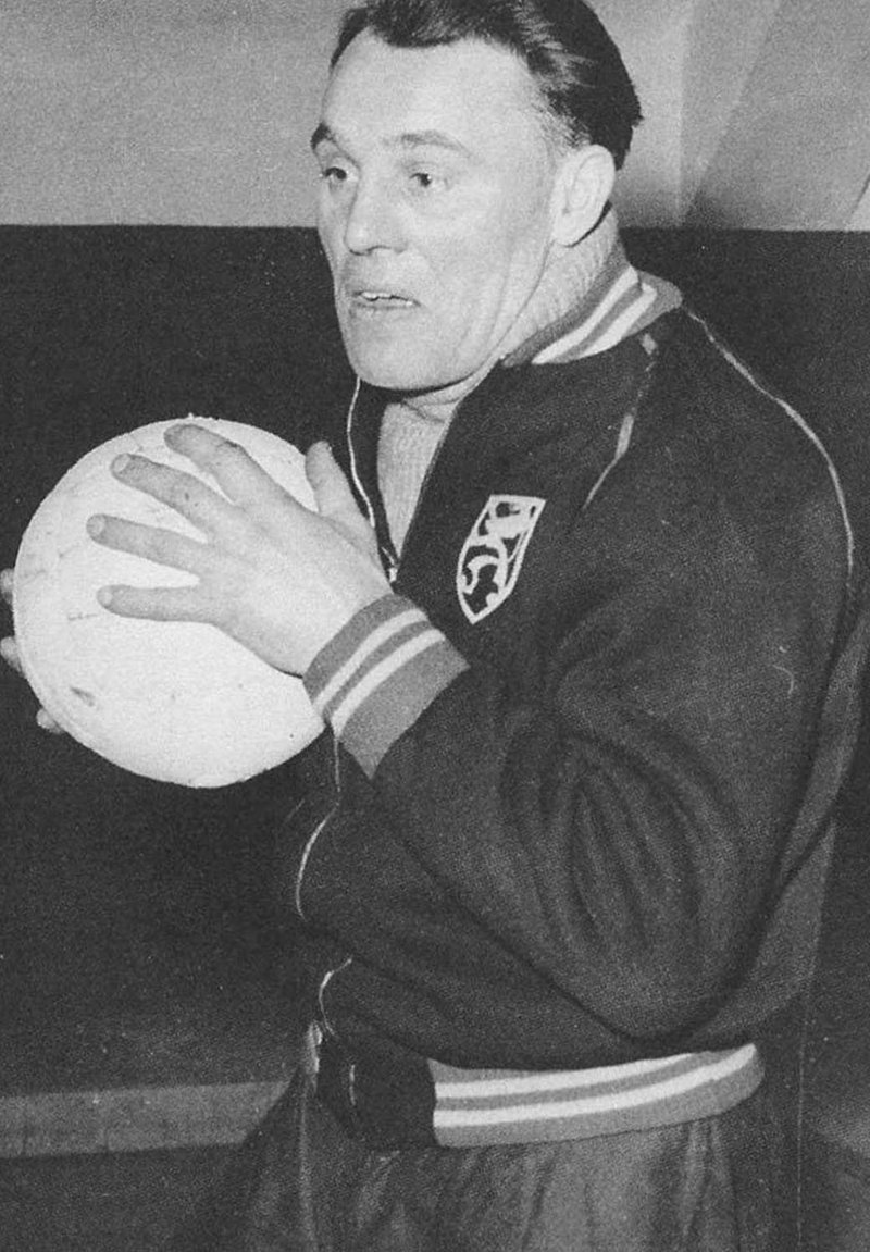 Zwart-wit afbeelding van een man die een textuurbal vasthoudt. Hij draagt een donkere jas met een logo en gestreepte accenten. Hij heeft kort haar.