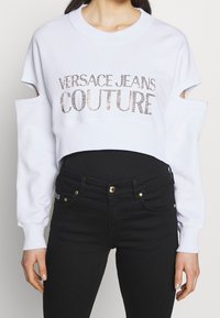 Vit kortärmad sweatshirt med utskurna axlar, med "VERSACE JEANS COUTURE" i skimrande silverbokstäver. Kombinerad med svarta jeans.