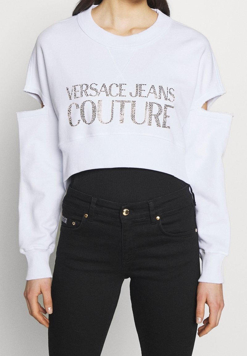 Vit kortärmad sweatshirt med utskurna axlar, med "VERSACE JEANS COUTURE" i skimrande silverbokstäver. Kombinerad med svarta jeans.