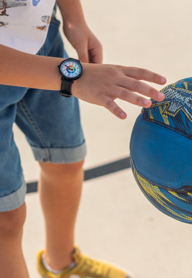 Montre noire avec un cadran coloré, shorts en jean et baskets jaunes, tendant la main vers un ballon de basket bleu avec des accents jaunes et gris.