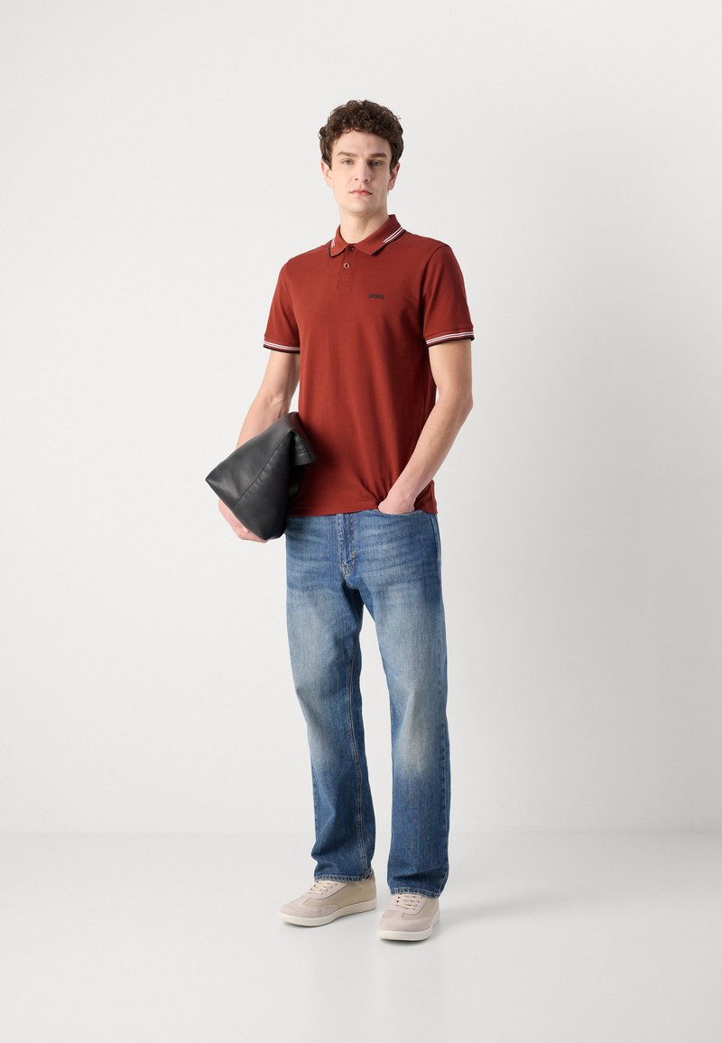 Polo rouge avec bordures blanches et bleu marine, logo noir ; jeans bleus ; tenant une pochette noire ; portant des baskets beiges ; debout sur un fond uni.