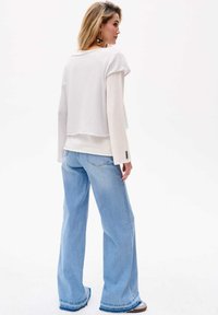 Weißer, verkürzter Sweatshirt mit kurzen Ärmeln, über einem langärmeligen Oberteil getragen, kombiniert mit hellblauen, weiten Jeans mit fransendem Saum.