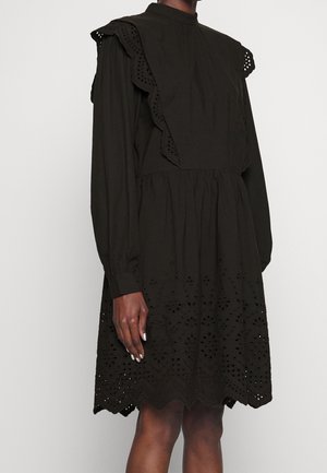 Robe noire à manches longues avec des détails en dentelle ajourée sur la jupe et des accents à volants sur les épaules portée par une personne à la peau de ton moyen-foncé.