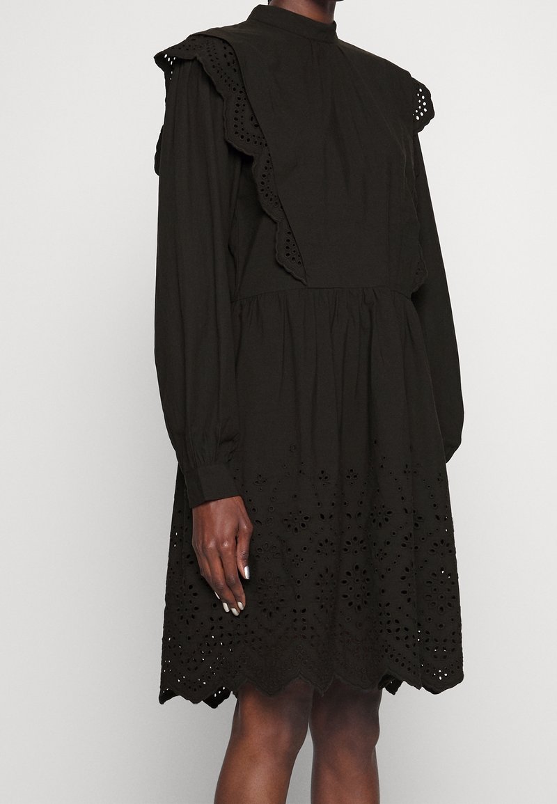 Robe noire à manches longues avec des détails en dentelle ajourée sur la jupe et des accents à volants sur les épaules portée par une personne à la peau de ton moyen-foncé.