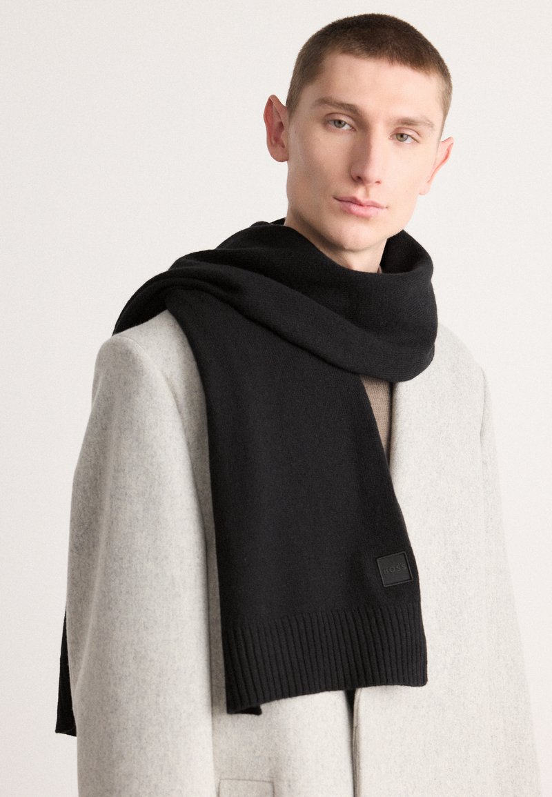 Black wool scarf with a ribbed hem, draped over a light gray coat. Features a branded tag on the end. Soft texture and long length.
