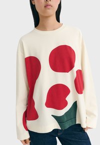 Woman wearing cream long-sleeve shirt with large abstract red and green shapes, paired with dark blue jeans.