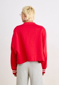 Red cropped sweatshirt with a high collar, dropped shoulders, and white striped cuffs, paired with gray sweatpants. Smooth texture.