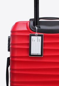 Valigia rigida rossa con superficie testurizzata, dettagli neri, manico telescopico e supporto per etichetta trasparente per l'identificazione.
