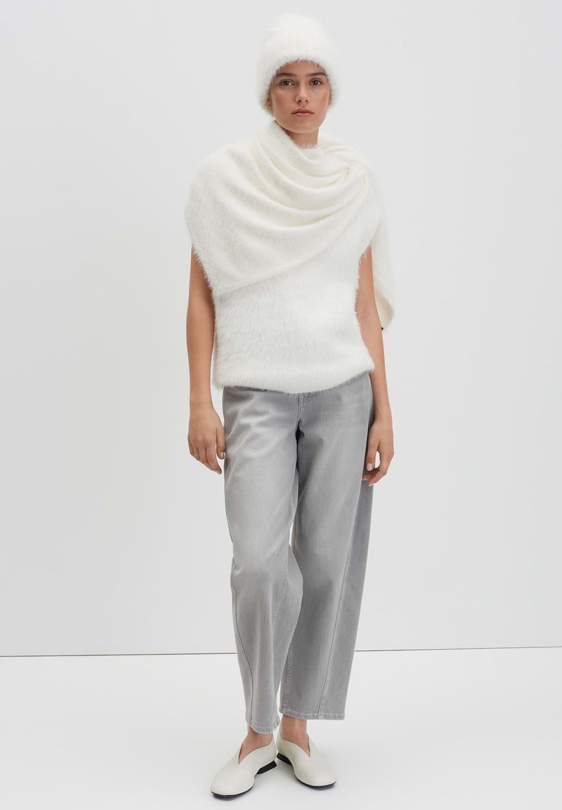 Woman wearing a white fuzzy hat, matching textured shawl and sleeveless top, gray wide-leg jeans, and white slip-on shoes against a white background.