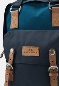 Batoh v námořnické modři a azurové látce s hnědými koženými popruhy a akcenty. Obsahuje nášivku s logem, stříbrné kovové doplňky a texturovaný povrch.