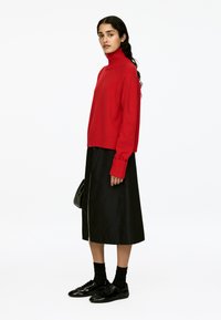 Frau mit langen dunklen Haaren, die einen roten Rollkragenpullover, einen schwarzen Midi-Rock mit Frontreißverschluss, schwarze Socken und glänzende schwarze Schuhe trägt und eine schwarze Tasche hält.